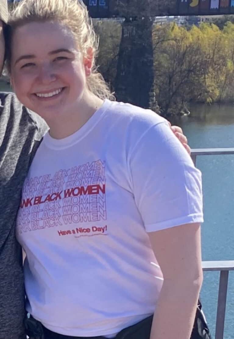 A smiling person with blond hair in a ponytail, wearing a "Thank Black Women" T-shirt, stands outdoors near water and trees—radiating fitness and positivity like an online fitness coach.